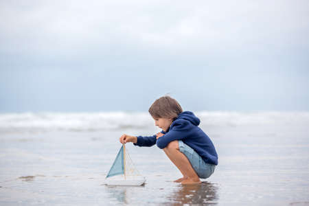 Child plays with sand on beach. Cute preschool boy with toy ship on beach. Stormy seaside sgore and kid playing on summer holidayの写真素材