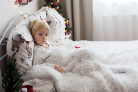 Beautiful blond toddler child, sick boy with fever, lying in bed on Christmas, coughing and having...