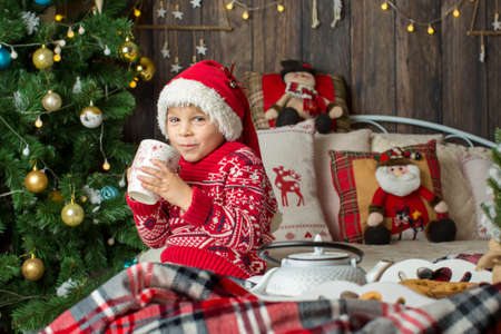 Cute toddler child, boy in a christmas outfut, playing in a wooden cabin on Christmas, derocation around him. Child reading book and drinking teaの写真素材