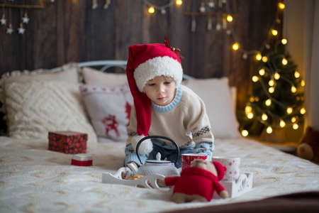 Cute toddler child, boy in a christmas outfut, playing in a wooden cabin on Christmas, derocation around him. Child reading book and drinking teaの写真素材