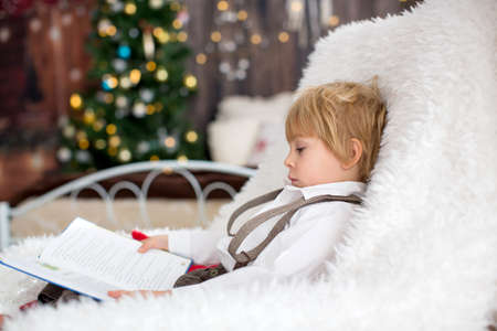 Adorable toddler child, sitting in cozy armchair with soft blanket, reading a book, christmas decoration around himの写真素材