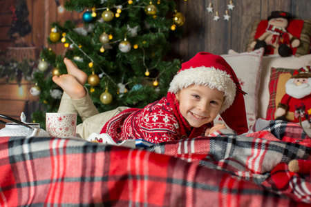 Cute toddler child, boy in a Christmas outfit, playing in a wooden cabin on Christmas, decoration around him. Child reading book and drinking teaの写真素材