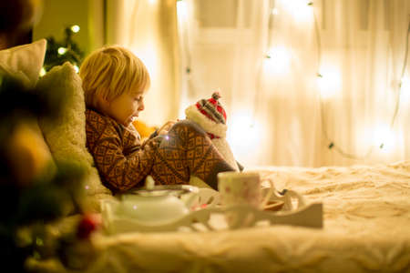 Cozy christmas atmosphere at home, child sitting on the bed, reading a book, enjoying christmas ligths and warm cup of teaの写真素材
