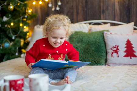 Cute toddler child, curly blond girl in a Christmas outfit, playing in a wooden cabin on Christmas, decoration around her. Child reading book and drinking teaの写真素材