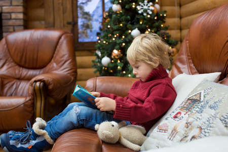 Blond toddler child, reading book and holding presents in a cozy cottage in the mountains, Christmas timeの写真素材