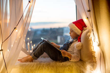 Toddler child, cute blond boy, sitting on the window, reading book, christmas lights aroundhimの写真素材