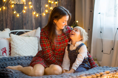 Toddler child hugging mother in bed, cute blond boy and mom embracing, christmas lights around themの写真素材