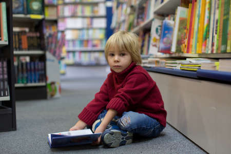 Cute blond toddler child, boy, reading book in a book store, wintertimeの写真素材