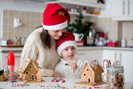 Blond toddler child and mom, cute boy and mother, decorating Christmas ginger bread house at home