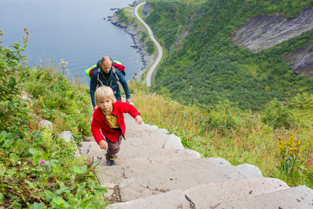 Children, toddler, brothers and father, climbing the stairs to Reinebringen, trekking path leading to the top of the mountain, where one can see Reine village and many fjords from aboveの写真素材