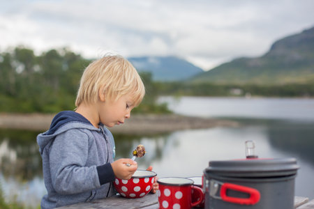 Happy child, eating on a bench on a rest stop along the road, mom cooking on small camping cooker and having family lunch on the roadの写真素材