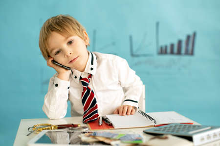 Good looking, smart casual dressed child, blond boy with tie, working on tablet, taking notes and talking on the phone, business concept and ideaの写真素材