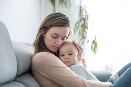 Young mother, holding her sick toddler boy, hugging him at home, sunny living room