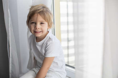 Cute happy toddler boy, child in white cloths, smiling at camera happily, playing next to a window with white curtainsの写真素材