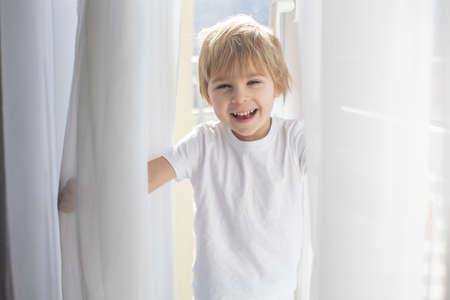 Cute happy toddler boy, child in white cloths, smiling at camera happily, playing next to a window...
