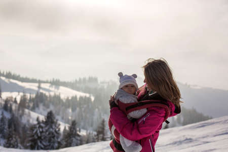 Young mother, carrying her baby boy in sling, climbing on peak wintertime, Austrian Apls, beautiful breath taking view from top of mountainの写真素材