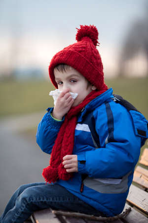 Little boy, sneezing and blowing his nose outdoor on a sunny winter day, sitting on a benchの写真素材