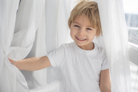 Cute happy toddler boy, child in white cloths, smiling at camera happily, playing next to a window with white curtainsの写真素材
