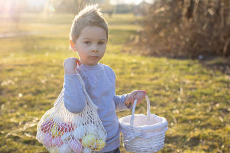 Cute toddler boy, gathering easter colorful eggs in a basket in the park, springtimeの写真素材