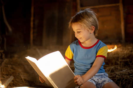 Amazing little blond child, boy, reading a book in a mystic cozy room filled with light from a small windowの写真素材