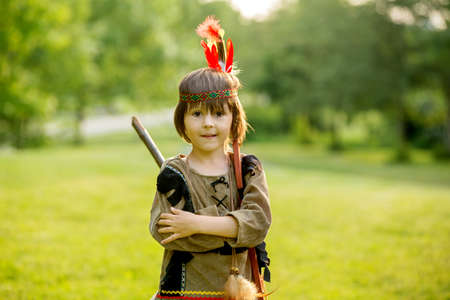 Cute portrait of native american boy with costumes, playing outdoor in the park with bow, arrows and hatchet on sunset, summertimeの写真素材