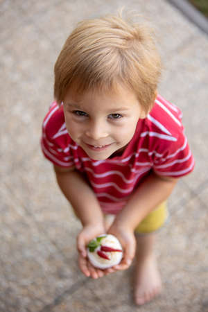 Cute child, boy, eating small cupcake of Pavlova desert, light egg and sugar desert with cream and strawberries, enjoyingの写真素材