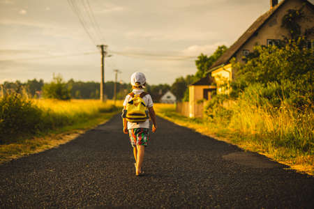 Beautiful child, boy with backpak walking on a little rural path on sunset, after rain, summertimeの写真素材