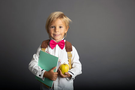 Cute preschool blond child, boy, holding books and notebook, apple, wearing glasses, ready to go to...