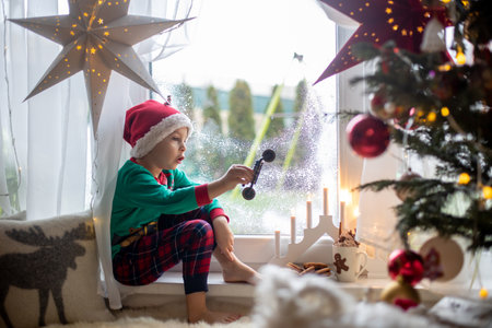 Toddler child, cute blond boy, sitting on the window in pajama, looking out for Satna Claus,, eating cookies,  Christmas lights around himの写真素材