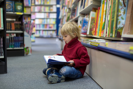 Cute blond toddler child, boy, reading book in a book store, wintertime