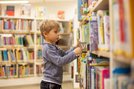 Adorable little child, boy, sitting in library, reading book and choosing what to lend, kid in book...