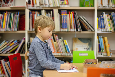 Adorable little child, boy, sitting in library, reading book and choosing what to lend, kid in book...