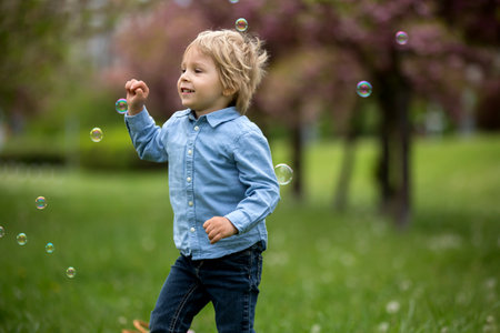 Blond toddler child, cute boy in casual clothing, playing with soap bubbles in the park, running...