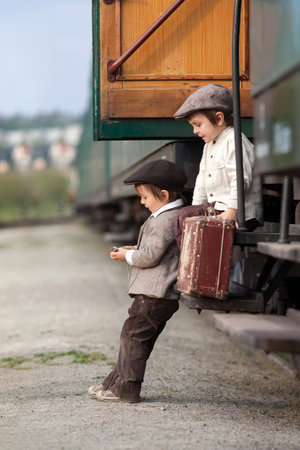 Two boys, dressed in vintage clothing and hat, with suitcase, on a railway stationの写真素材