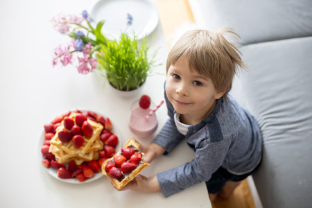 Sweet preschool child, boy, eating belgian waffle with strawberries and chocolate at home and driking smootieの写真素材