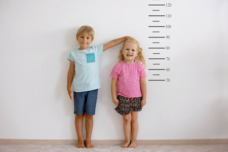 Little children, blond boy and girl, measuring height against wall in room, comparing themselves