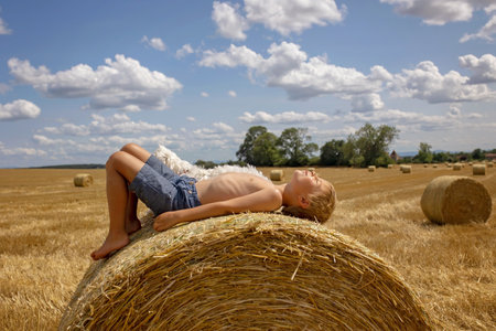 Beautiful blond child, boy, lying on a haystack in the field. Amazing landscape, rural scene with clouds, tree and empty road summertime, fields of haystack next to the road, summer in Portugalの写真素材