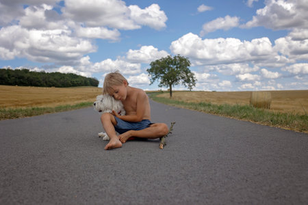 Beautiful blond child, boy, walking on rural road with his sweet little maltese pet dog. Amazing landscape, rural scene with clouds, tree and empty road summertime, fields of haystack next to the road, summerの写真素材