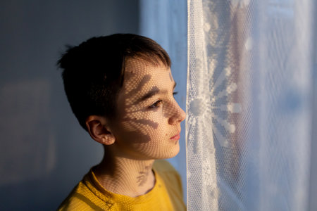Artistic portrait of a teenage boy, standing next to a window, shadows from curtains falling on his face, making interesting shadows, art portraitの写真素材