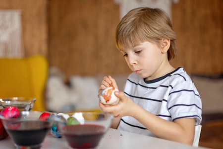 Beatiful blond child, boy, coloring and painting eggs for Easter at home, preparing for the holidays