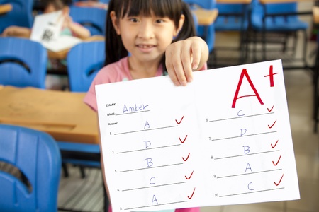 smiling little girl showing exam paper with a plus in the classroomの写真素材