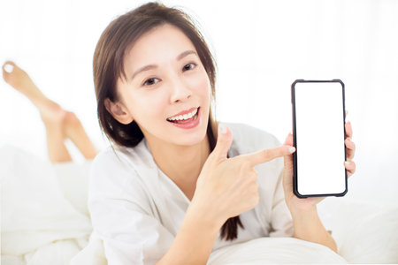 happy young woman showing smart phone while lying on the bedの写真素材