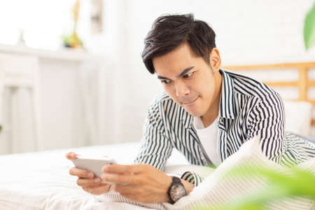 young man wearing casual clothes at home, lying in bed using laptop to surf the Internet,...