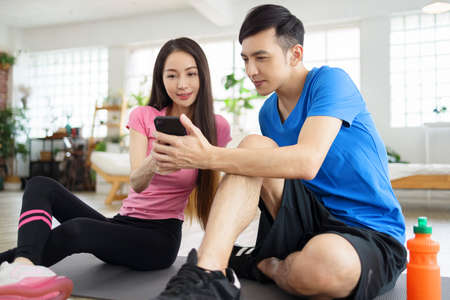 Athletic happy couple sitting on fitness mat, watching the mobile phone.