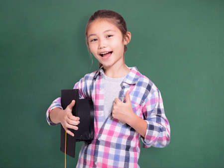 Happy student girl holding graduation cap and showing thumb up.Isolated on green chalkboard background.の写真素材