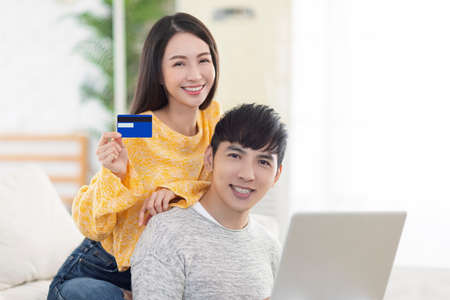 Young couple showing credit card and using laptop in the living room.