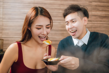 Young asian man feeding happy girlfriend with tasty food in restaurantの写真素材