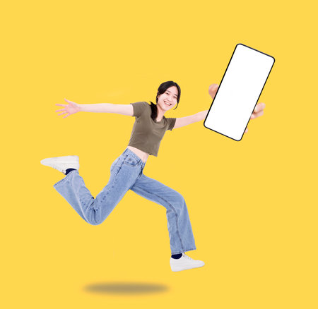 Happy girl student showing the blank screen mobile phone jump and run wear t-shirt denim isolated yellow backgroundの写真素材