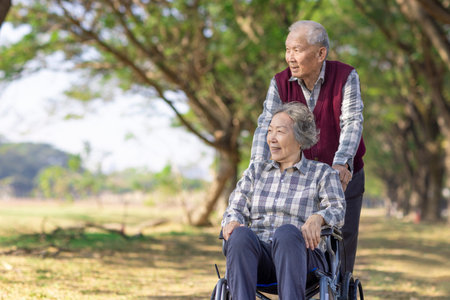 Happy Asian senior couple enjoys park stroll. Elderly man pushes wife in wheelchair