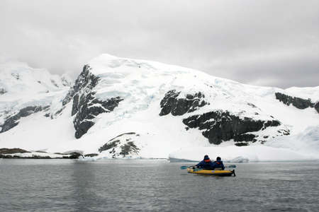Two people in yellow kayak in Antarctica. Shore under snow and cloudy sky.の写真素材